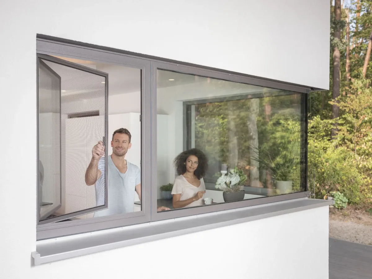 Mann öffnet ein Fenster mit Fliegengitter, Frau steht im Raum dahinter – Insektenschutz Fenster nach Mass von GP Insektenschutz