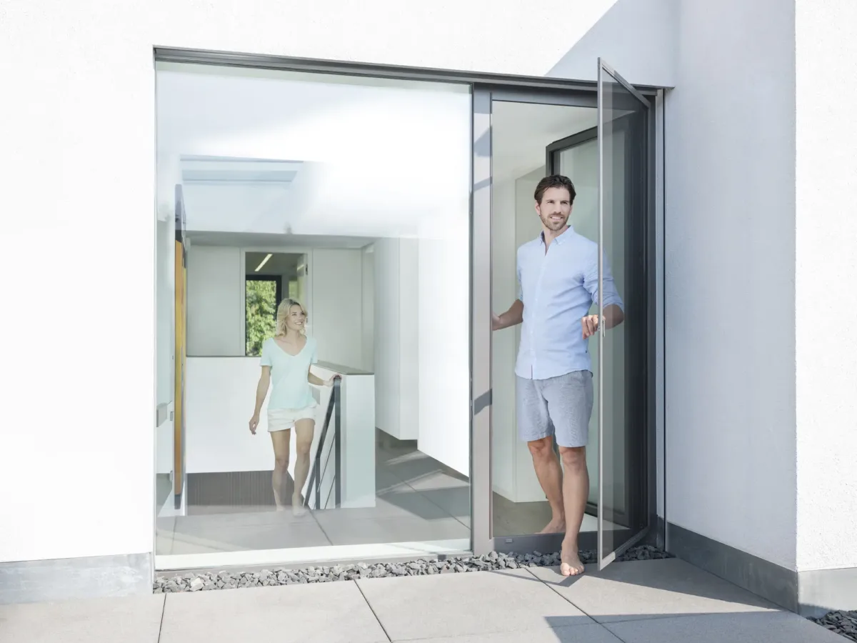 Mann tritt durch eine Insektenschutz Tür nach Mass auf die Terrasse, im Hintergrund steht eine Frau im Haus – Insektenschutz Produkte von GP Insektenschutz für moderne Balkontüren