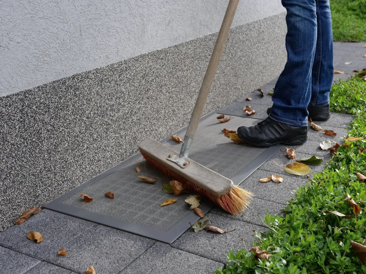 Mann kehrt begehbare Lichtschachtabdeckung mit Insektenschutz vor Kellerfenster