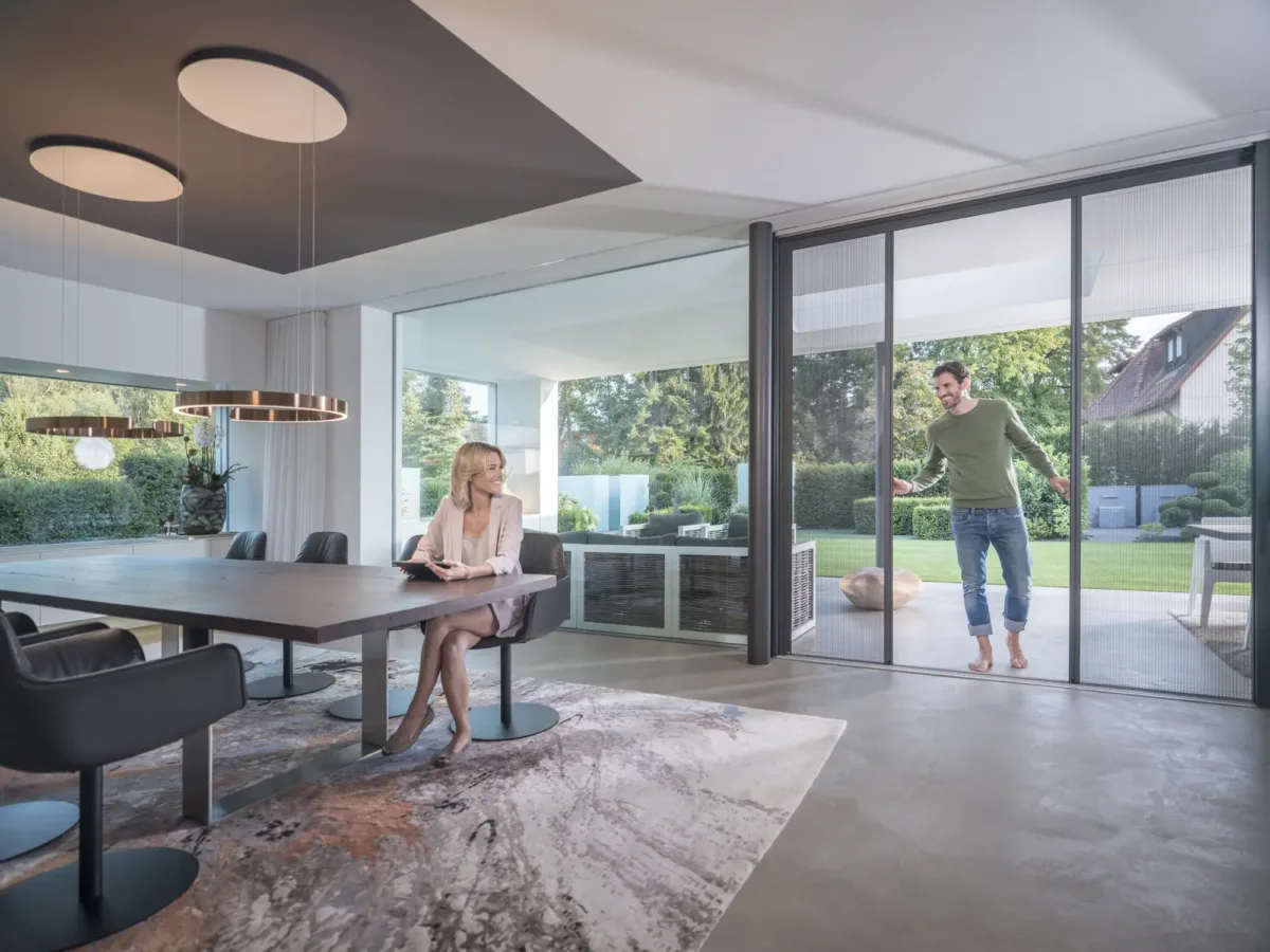 Frau schiebt eine seitlich laufende Plissee Insektenschutztür vor der Terrassentür, drahtseilgeführtes System mit Rollenlauf